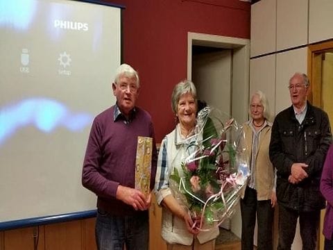 Vier Menschen bei einer kleinen Feier vor einer Leinwand, am rechten Rand angeschnitten eine weitere Person; eine Frau hält einen großen Blumenstrauß, ein Mann ein Geschenk.