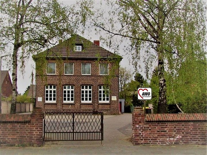 Die Begegnungsstätte Alte Schule: ein zweistöckiges Backsteinhaus mit Bogenfenstern hinter einer Mauer mit Gittertor, rechts das Schild „AWO Sinnersdorf“.